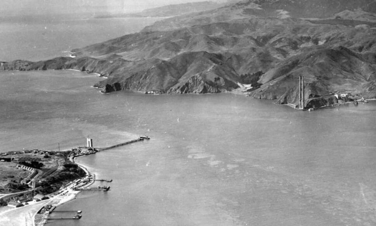 Aerial view of the San Francisco Bay with hills in the background and the early construction of the Golden Gate Bridge, showing only the two towers rising from the water.