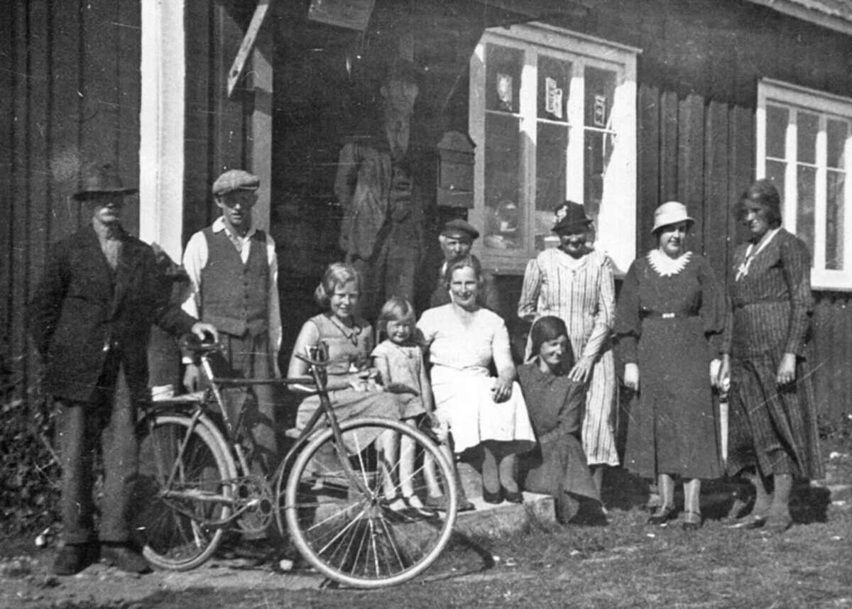A black-and-white photo of a group of people, including men, women, and children, standing and sitting outside a wooden building. A bicycle is in the foreground near the group. The image appears old and vintage.