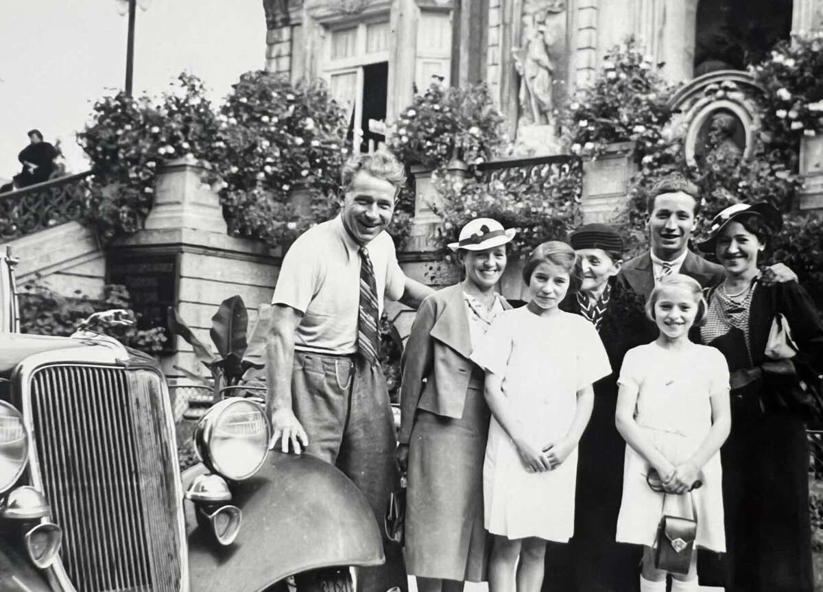 A black-and-white photo of six people posing and smiling in front of a vintage car and an ornate building with blooming flowers. Three adults stand behind three girls, all dressed in 1930s-style clothing and hats.