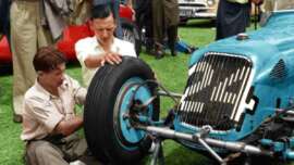 Two men work together to attach a large tire to the front wheel of a vintage blue race car, surrounded by other people and vehicles on a grassy area.