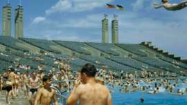 Crowded outdoor swimming pool with people relaxing and swimming. In the background, large empty stadium seating and two flagpoles with German flags are visible under a partly cloudy sky.