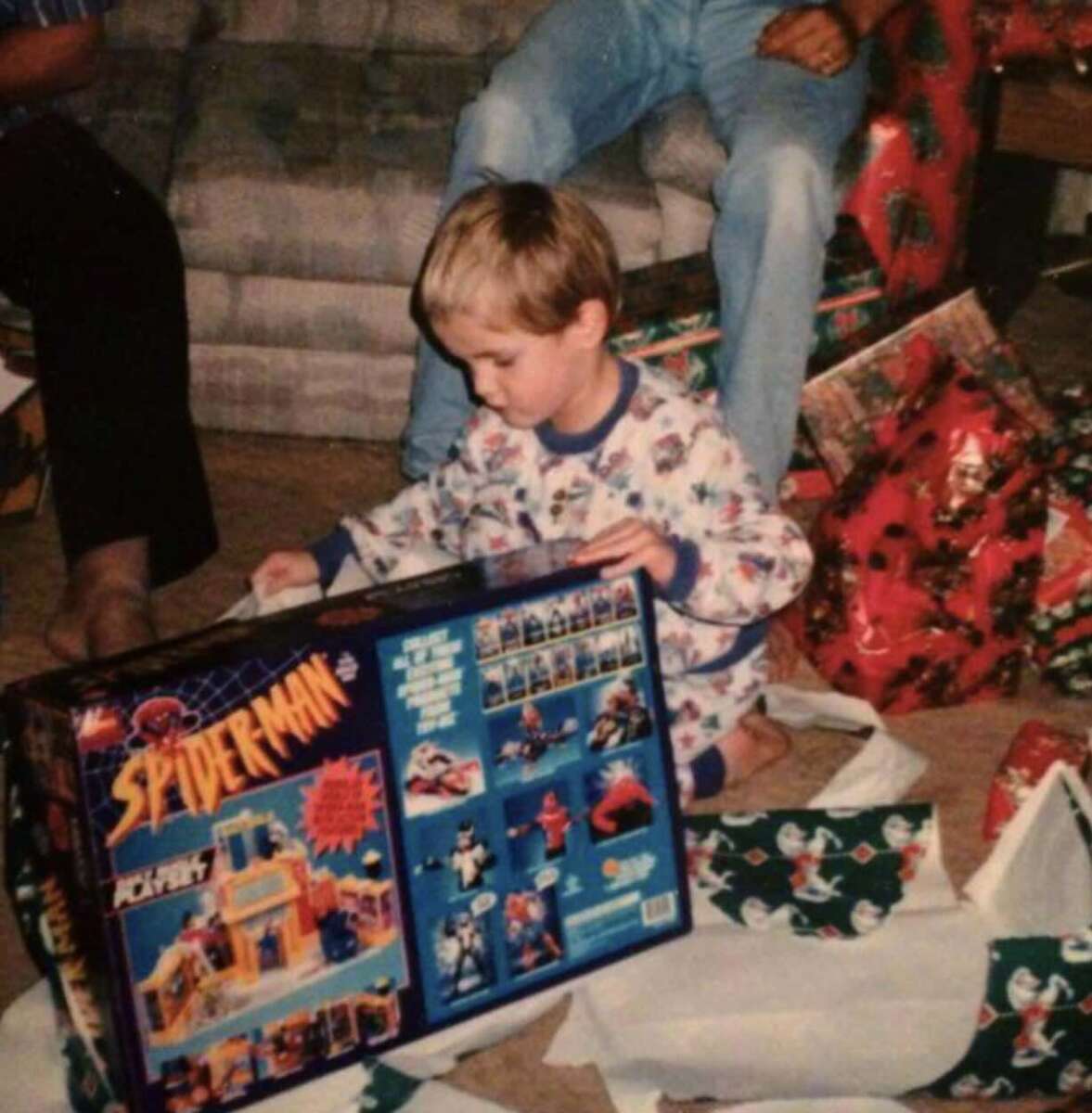 A young child in pajamas sits on the floor amid wrapping paper, excitedly holding a large Spider-Man toy set box in front of a sofa, with an adult’s legs visible behind him.