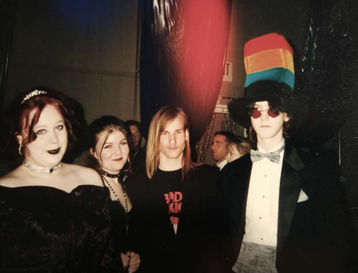 Four people pose together at an event. One wears a tiara and black lipstick, another has a necklace and dark makeup, a third wears a shirt with red text, and the fourth has a tall rainbow hat, round red glasses, and a bow tie.