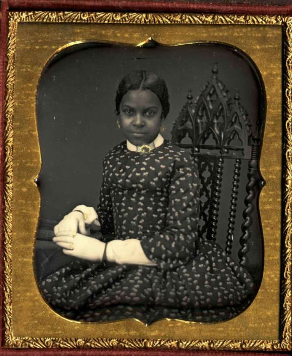A young Black girl wearing a patterned dress sits on a decorative chair, hands folded in her lap, gazing calmly at the camera. The photograph is in an ornate gold-colored frame.