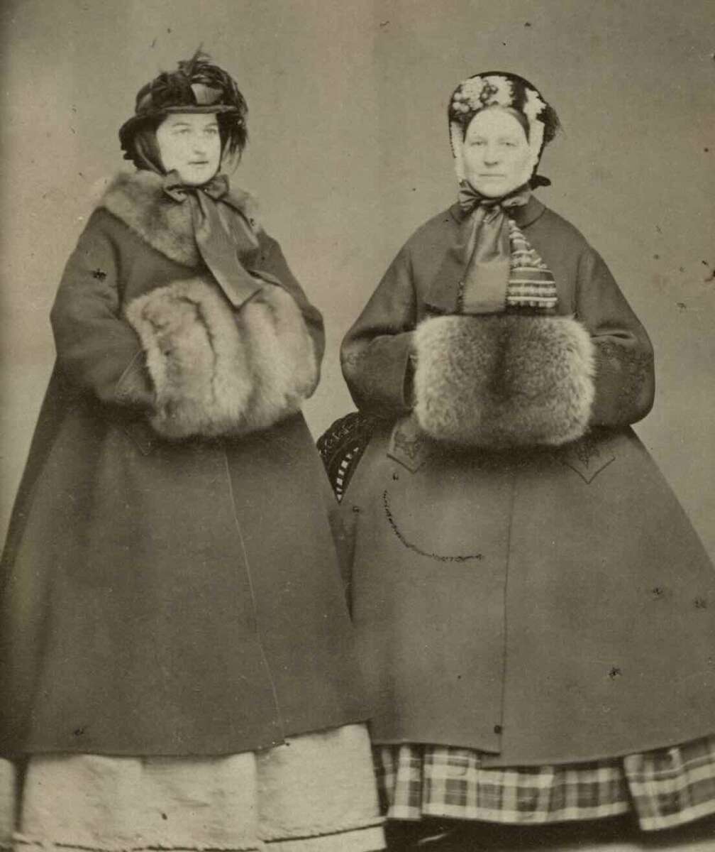 Two women in 19th-century winter clothing stand side by side, each wearing a cloak, a bonnet, and holding a large fur muff. Their skirts are full and patterned, and they face the camera with neutral expressions.