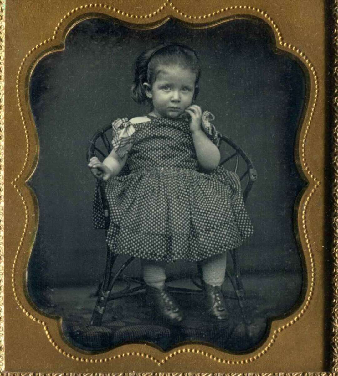 A young girl in a polka-dot dress sits on a small chair, looking slightly to the side with one hand near her face. The photo has an ornate, gold-colored frame and a vintage, sepia-tone appearance.