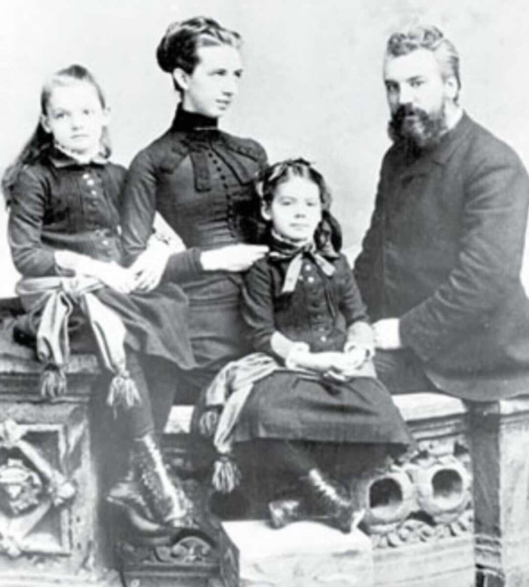 A vintage black-and-white photo of a family of four, with two young girls and two adults, all dressed in dark, formal Victorian-era clothing, seated and posing for a portrait.