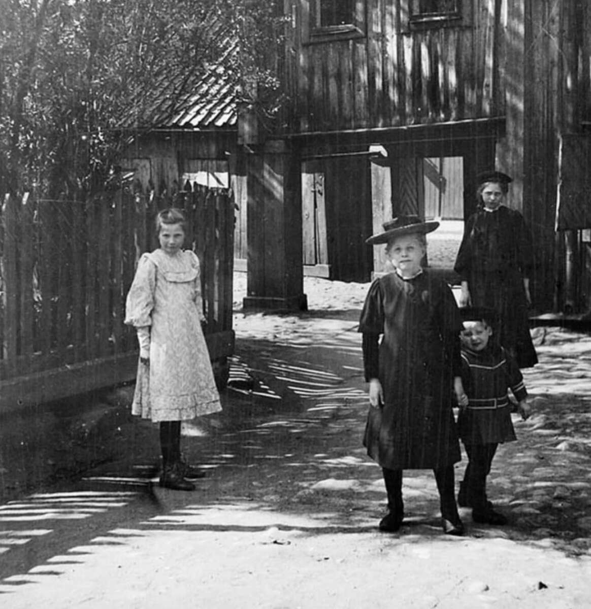 Four people, including two women and two children, stand in a sunlit yard with sandy ground, wooden fence, and old wooden buildings. The women and children wear early 20th-century clothing and hats.