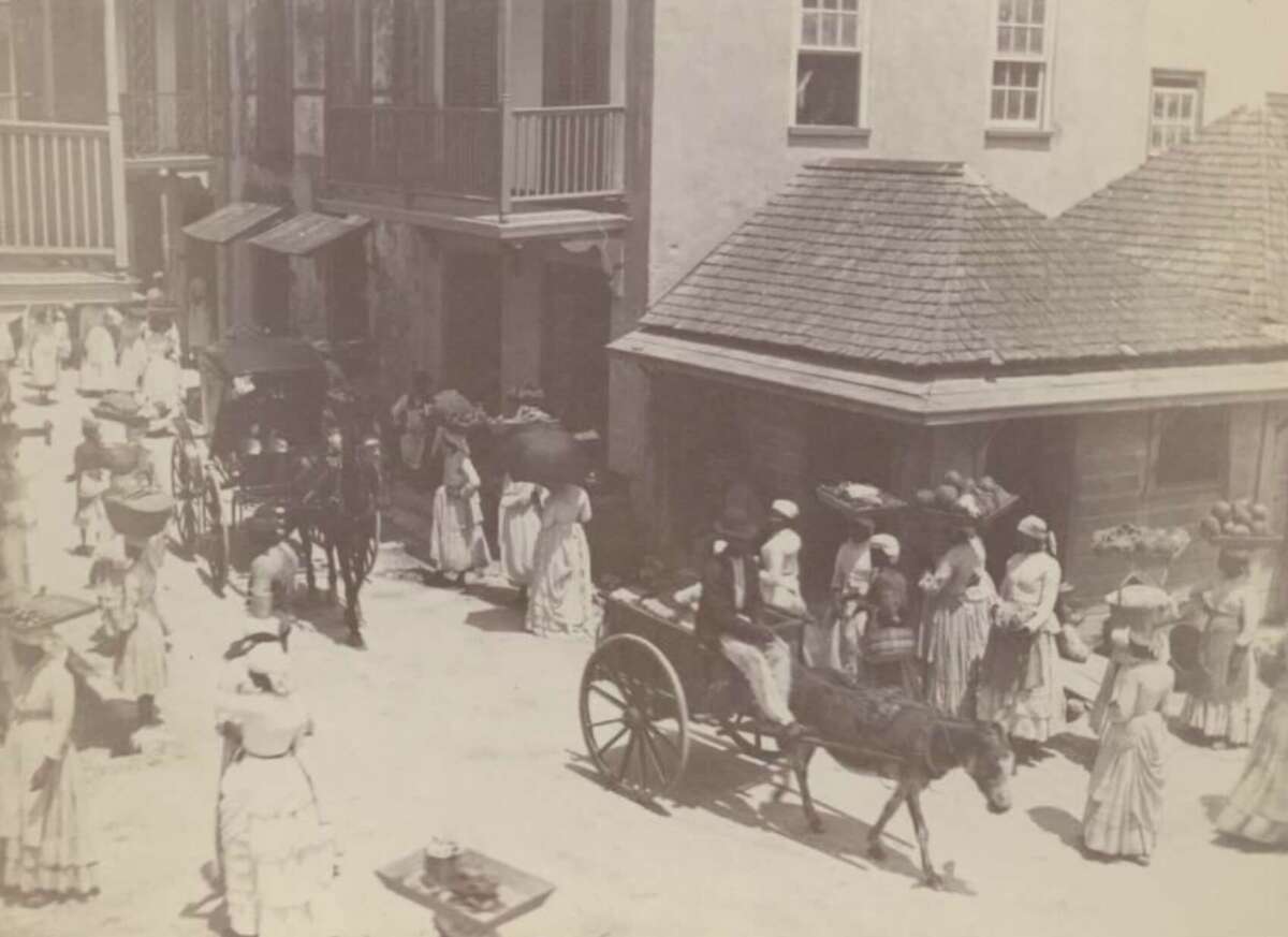 Black-and-white photo of a busy street scene with women in dresses and bonnets, horse-drawn carts, and buildings with wooden balconies. People are gathered on both sides, some appearing to sell goods.