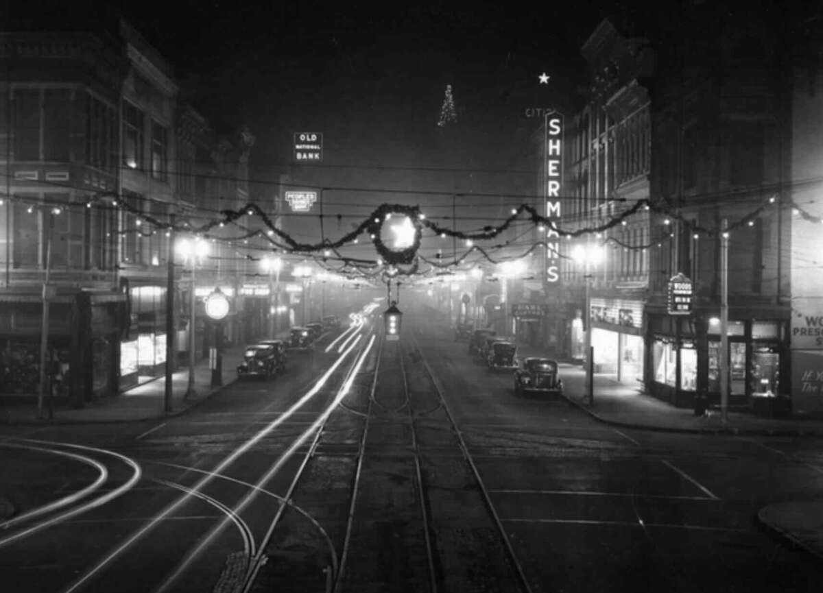 A black-and-white photo of a city street at night in the 1940s, decorated with garlands and lights. Old cars are parked along the sidewalks, and vintage shop signs, including "Sherman's," are illuminated.