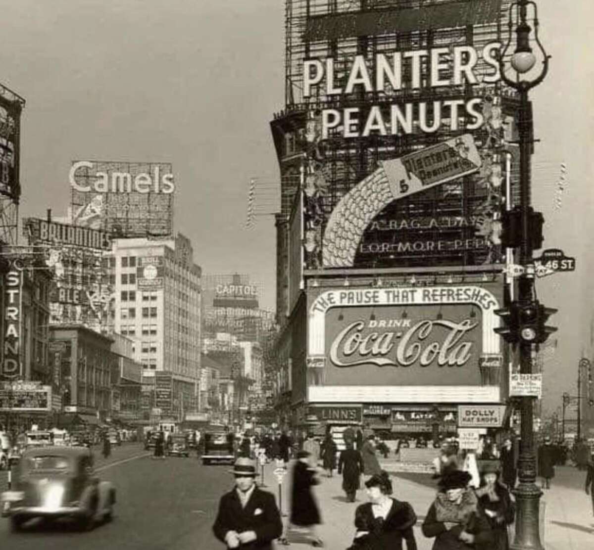A busy city street scene from the early 20th century, featuring vintage cars and pedestrians. Large neon advertisements for Planters Peanuts, Camels, and Coca-Cola dominate the buildings.