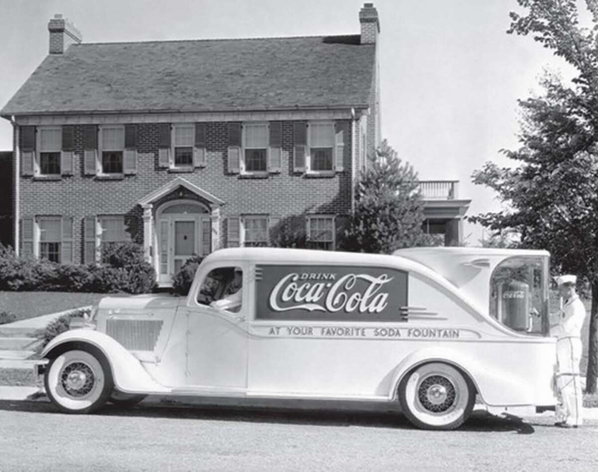 A vintage Coca-Cola delivery truck is parked in front of a large brick house. A person in a white uniform stands beside the truck, which features a large Coca-Cola logo and old-fashioned styling.