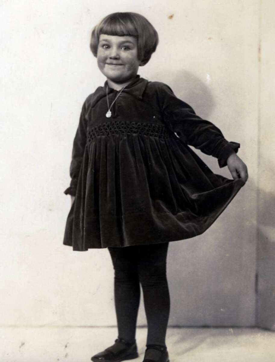 A young child with a short bob haircut smiles while holding out the skirt of a dark, long-sleeved dress. The child is wearing dark tights, shoes, and a necklace, standing against a plain background.