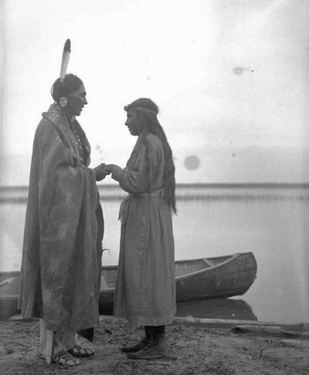 A man and a woman stand facing each other near a canoe by the water. The man wears traditional clothing and a feather in his hair; the woman has long hair and a dress. They appear to be holding hands or exchanging something.