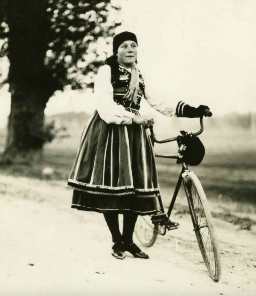 A woman in traditional dress stands on a dirt road, holding a vintage bicycle. Trees and open countryside are visible in the background. The photo appears old and in black and white.