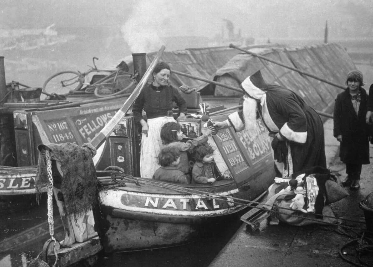 A person dressed as Santa Claus hands gifts to children on a canal boat, while a woman and another child stand nearby. The scene appears old-fashioned, with smoke and rustic boats in the background.