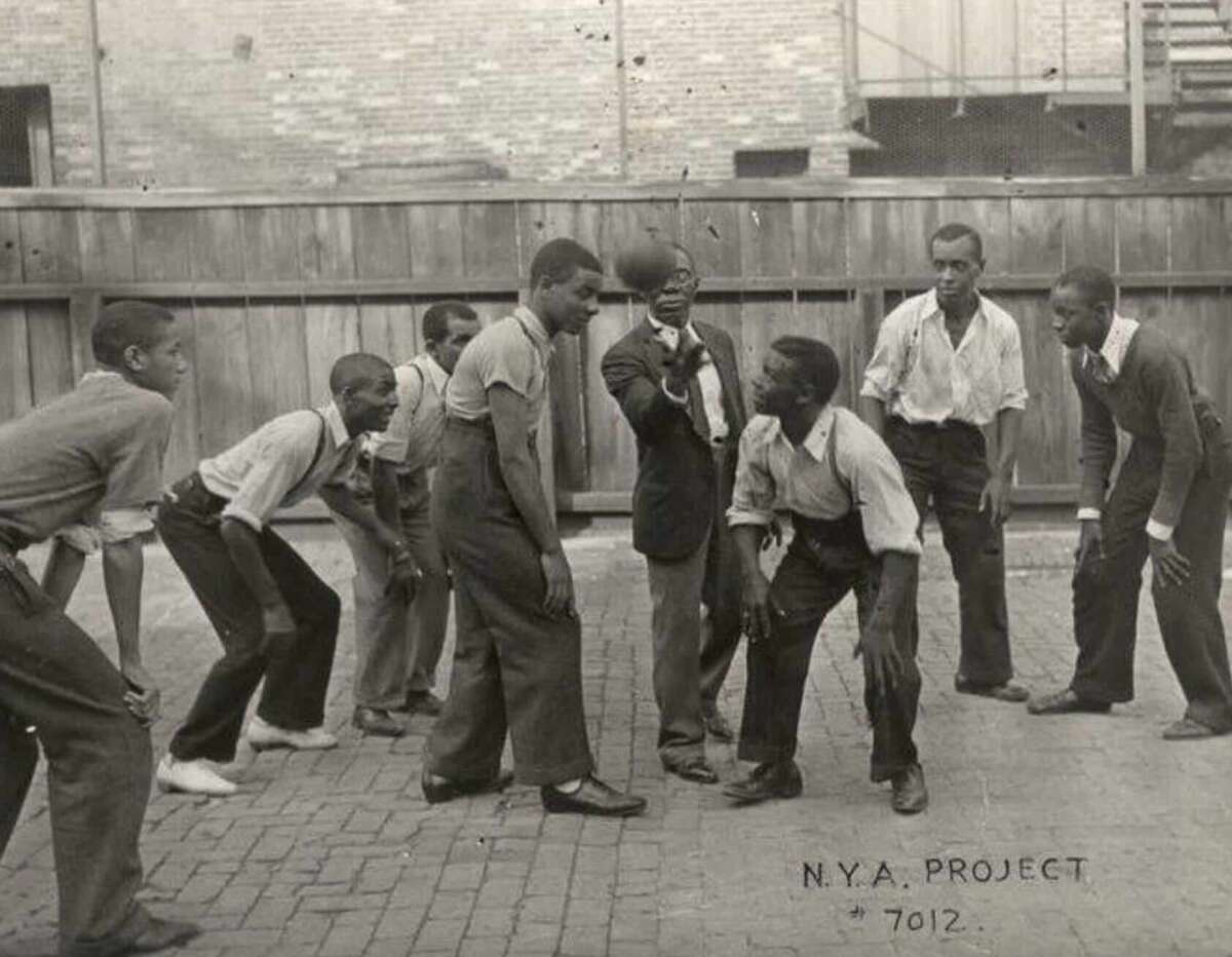 A group of young Black men play basketball outdoors on a paved court, with a man in a suit tossing the ball. A wooden fence and brick wall are in the background. "N.Y.A. PROJECT 7012" is written on the photo.
