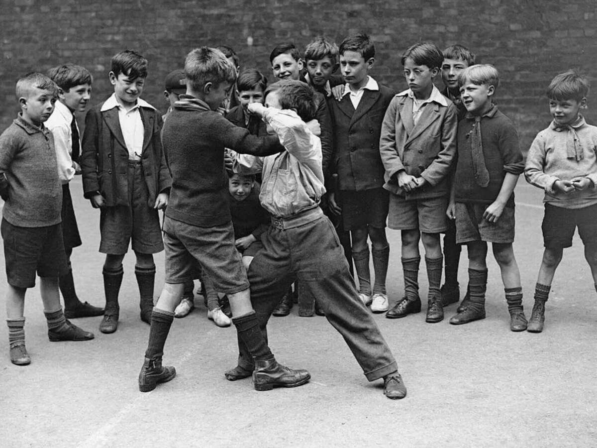 A group of boys in vintage clothing stand outside, watching two boys in the center playfully fighting or wrestling, while others look on with interest and curiosity.