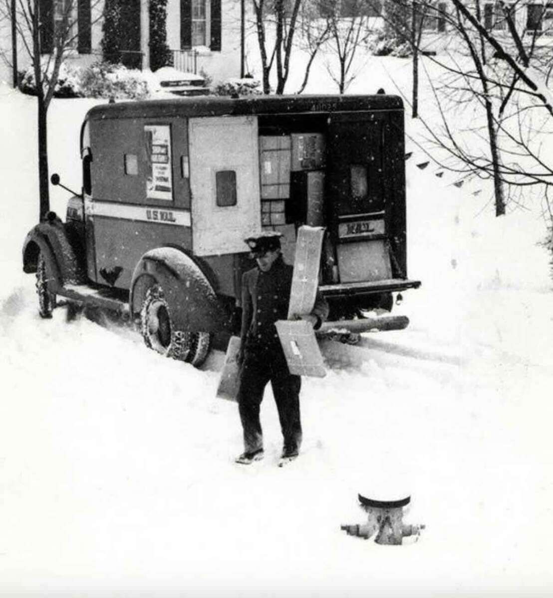 A postal worker carries packages through deep snow from a vintage U.S. Mail truck parked on a snowy residential street, with houses and bare trees in the background.