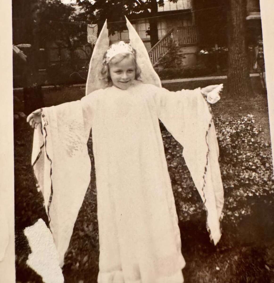 A young child in a white costume with wing-like sleeves and a halo-like headpiece poses with arms outstretched in a garden, in front of a house. The black-and-white photo has a vintage feel.