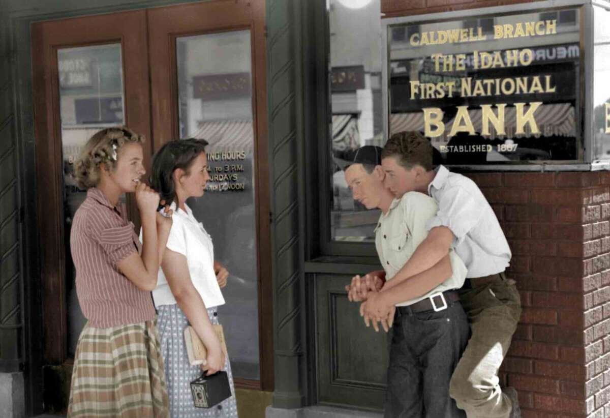 Two young women stand talking outside a brick bank building as two young men, one hugging the other from behind, stand nearby under a sign reading "The Idaho First National Bank.