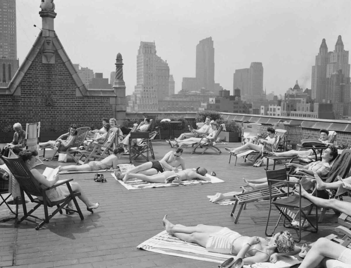 People sunbathe and relax on lounge chairs and towels on a rooftop in a city, surrounded by tall buildings, with a hazy skyline in the background. Most are in swimsuits, enjoying the sunny day.