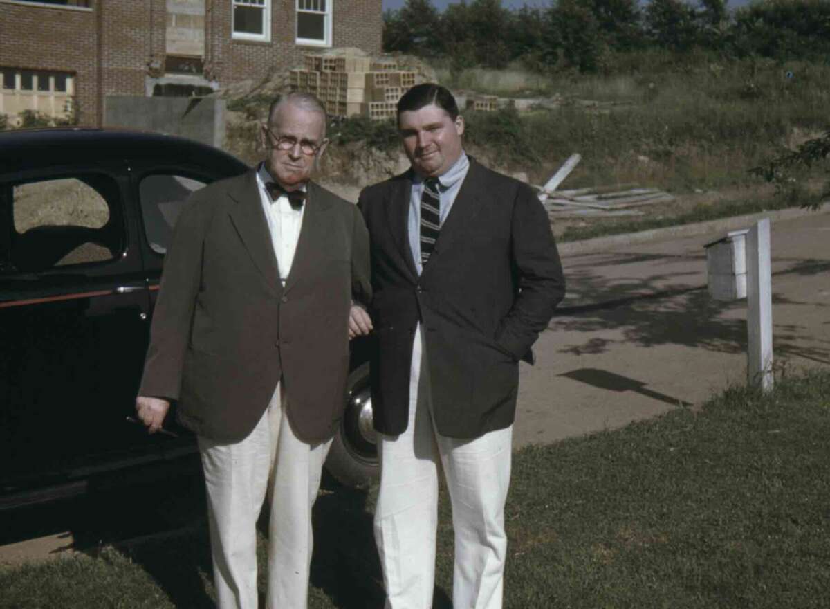 Two men in suits and ties stand outdoors beside a black car on a sunny day, with a building and construction materials visible in the background. Both men are smiling.