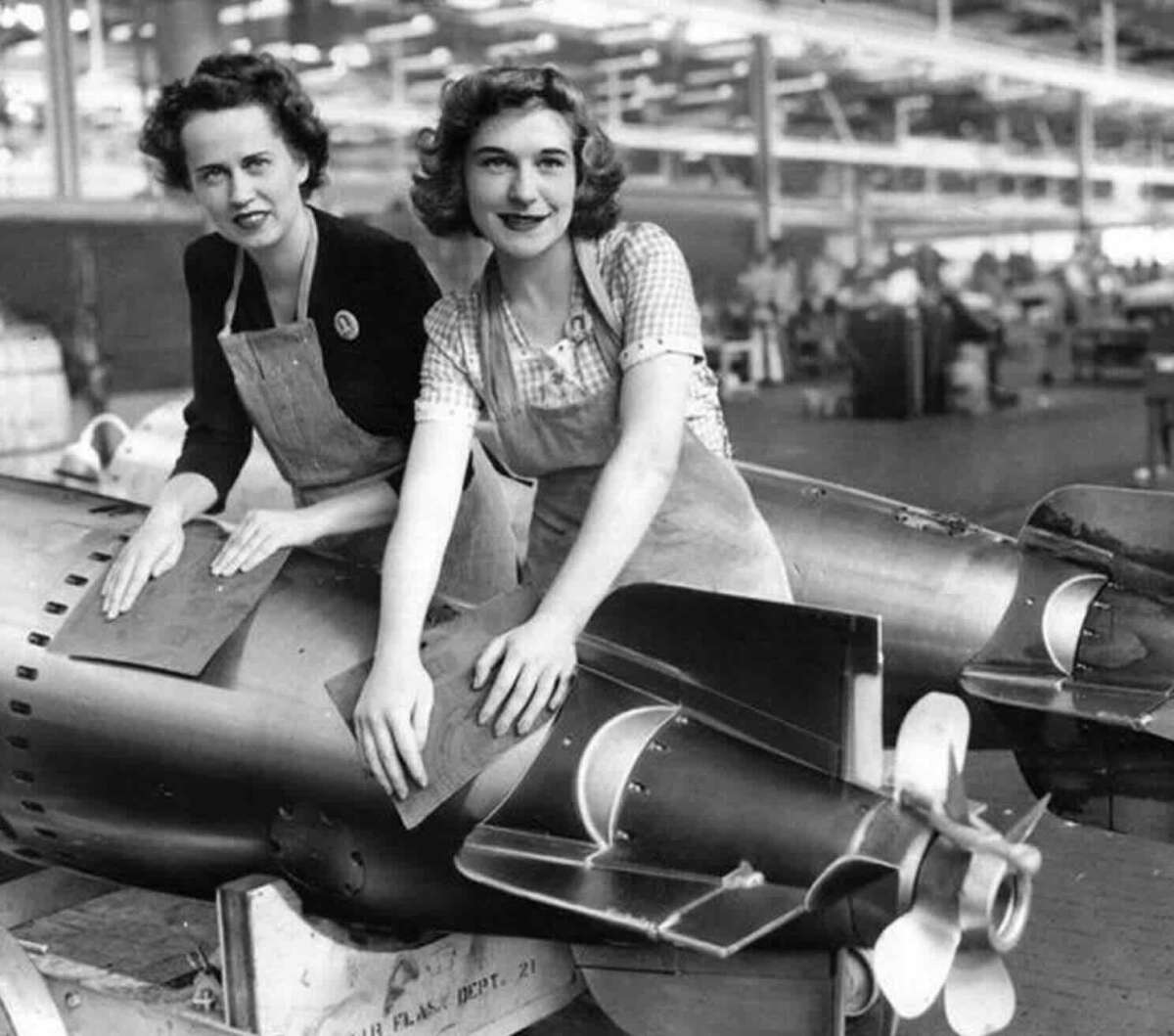 Two women in aprons work on assembling or inspecting a large bomb or missile in a factory setting, likely during World War II. They are smiling and appear focused on their tasks.