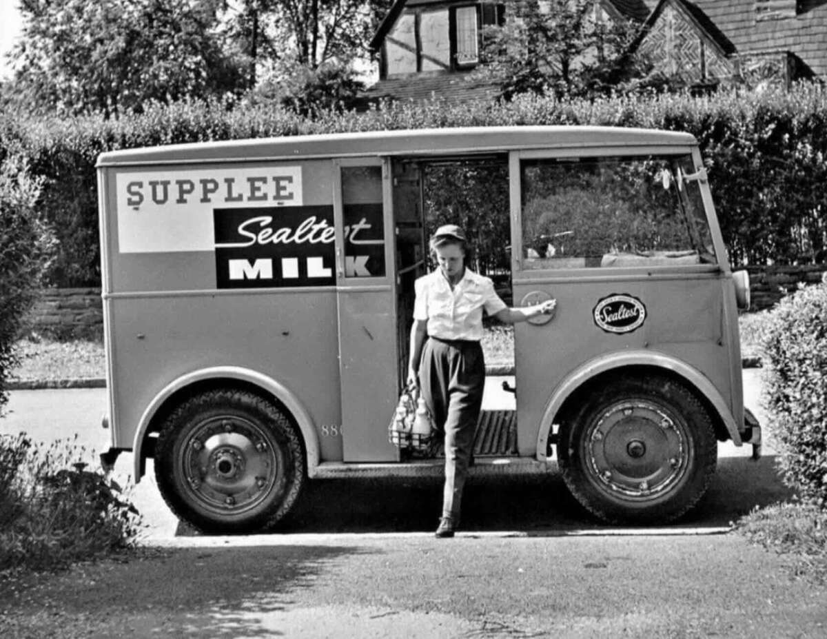 A woman stands beside an old-fashioned Supplee Sealtest Milk delivery truck, holding a metal milk carrier, in a suburban setting with trees and a house in the background.