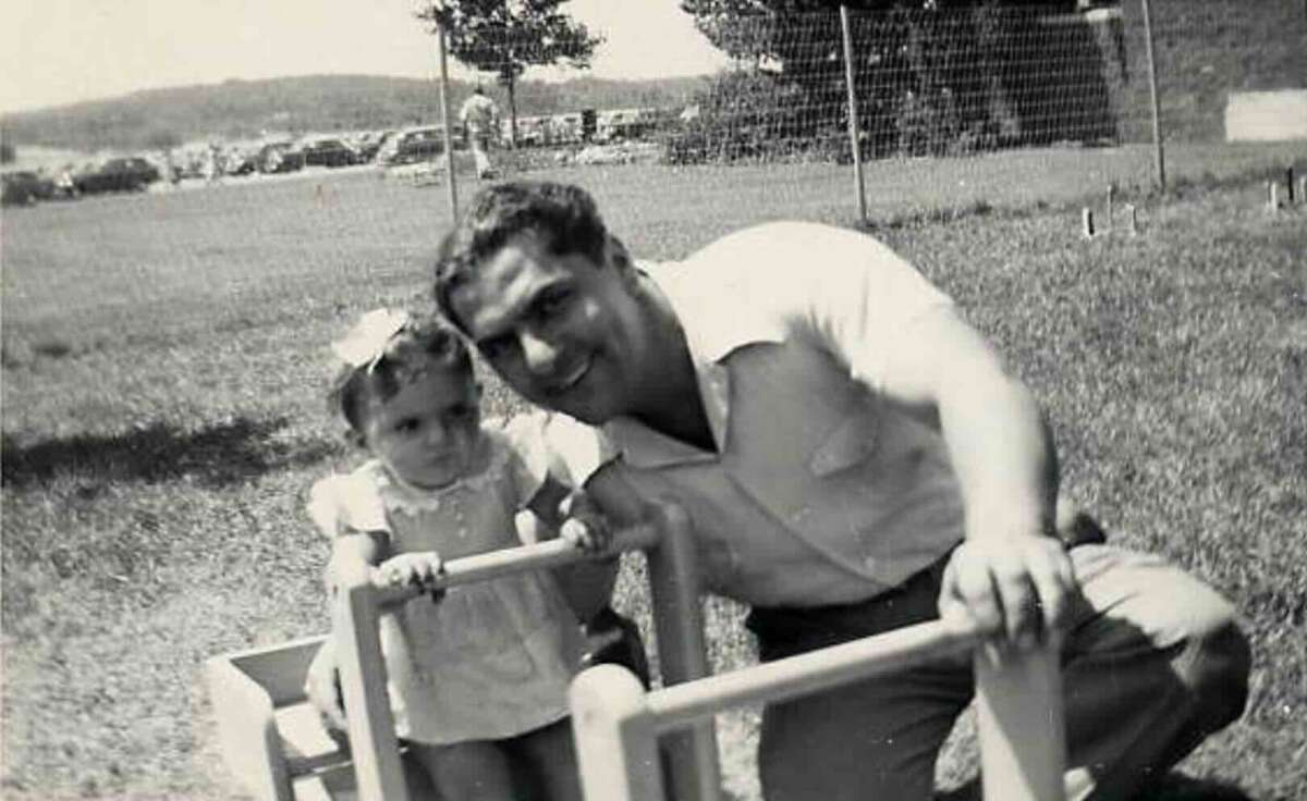 A man crouches beside a young girl holding onto playground equipment in a grassy outdoor area; both are smiling and posing for the black-and-white photo. Trees and a fence are visible in the background.