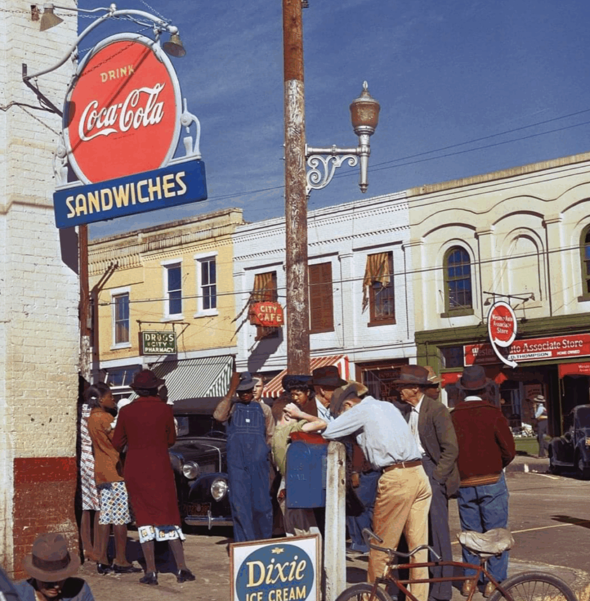 A group of people gather on a sunny street corner near a Coca-Cola and sandwiches sign, with vintage cars and colorful storefronts, evoking a mid-20th-century small-town setting.