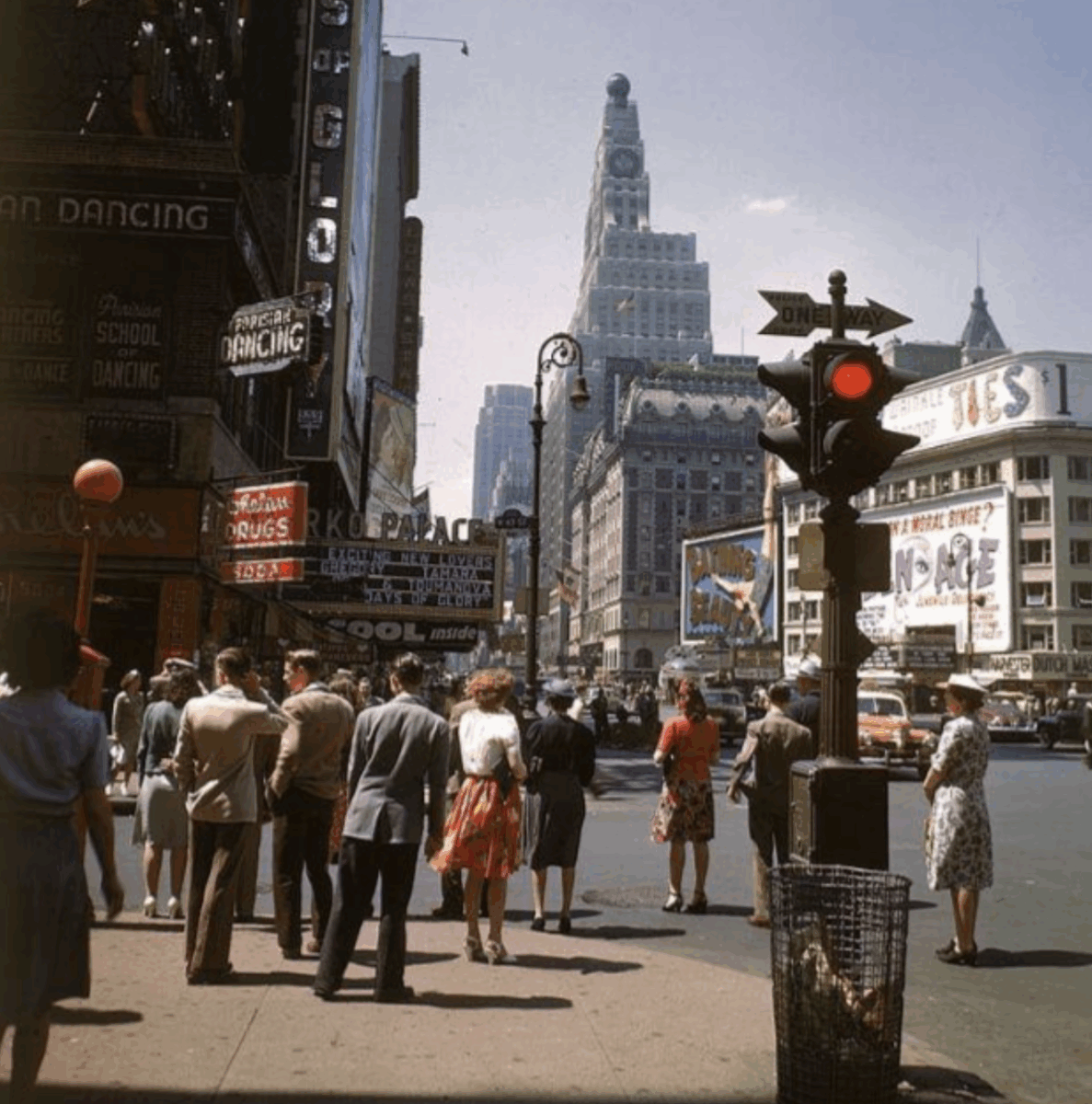 A busy city street scene from the 1940s with people waiting at a crosswalk, vintage cars, neon signs, and tall buildings under a blue sky. A traffic light shows red, and advertisements line the storefronts.
