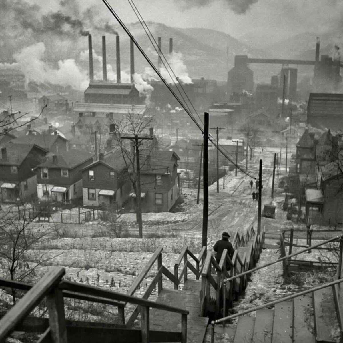 A black-and-white photo shows a person descending snowy wooden steps toward a town with houses and smoke billowing from multiple factory chimneys in the background, creating an industrial, wintry scene.