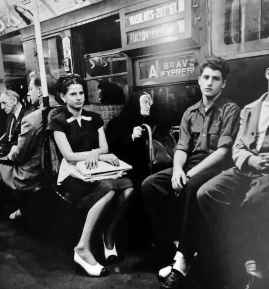 Black-and-white photo of people sitting quietly on a New York City subway train, including a young woman and man in the foreground, with vintage advertisements and signs visible above and behind them.