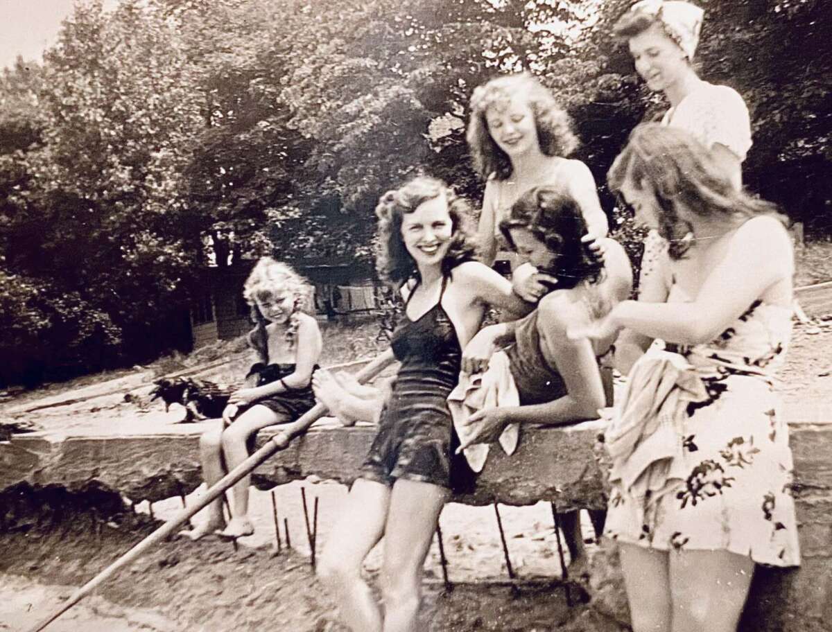 A vintage black-and-white photo of six young women in swimsuits and summer dresses posing and smiling together on a sandy beach, with trees in the background.