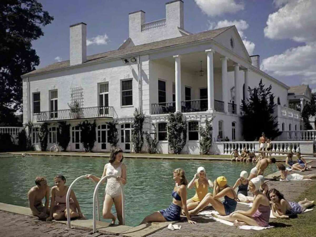 People lounge and socialize by a large outdoor pool in front of a grand, white mansion on a sunny day. Some wear swimsuits and swim caps, while others sit or stand near the pool’s edge, enjoying the relaxed atmosphere.