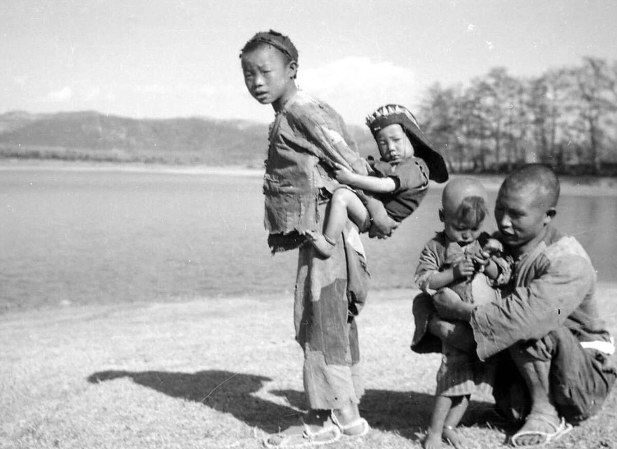 Four children in worn clothing stand by a lake; one child carries a baby on their back, while another crouches, helping a smaller child. The background features water, trees, and distant mountains.