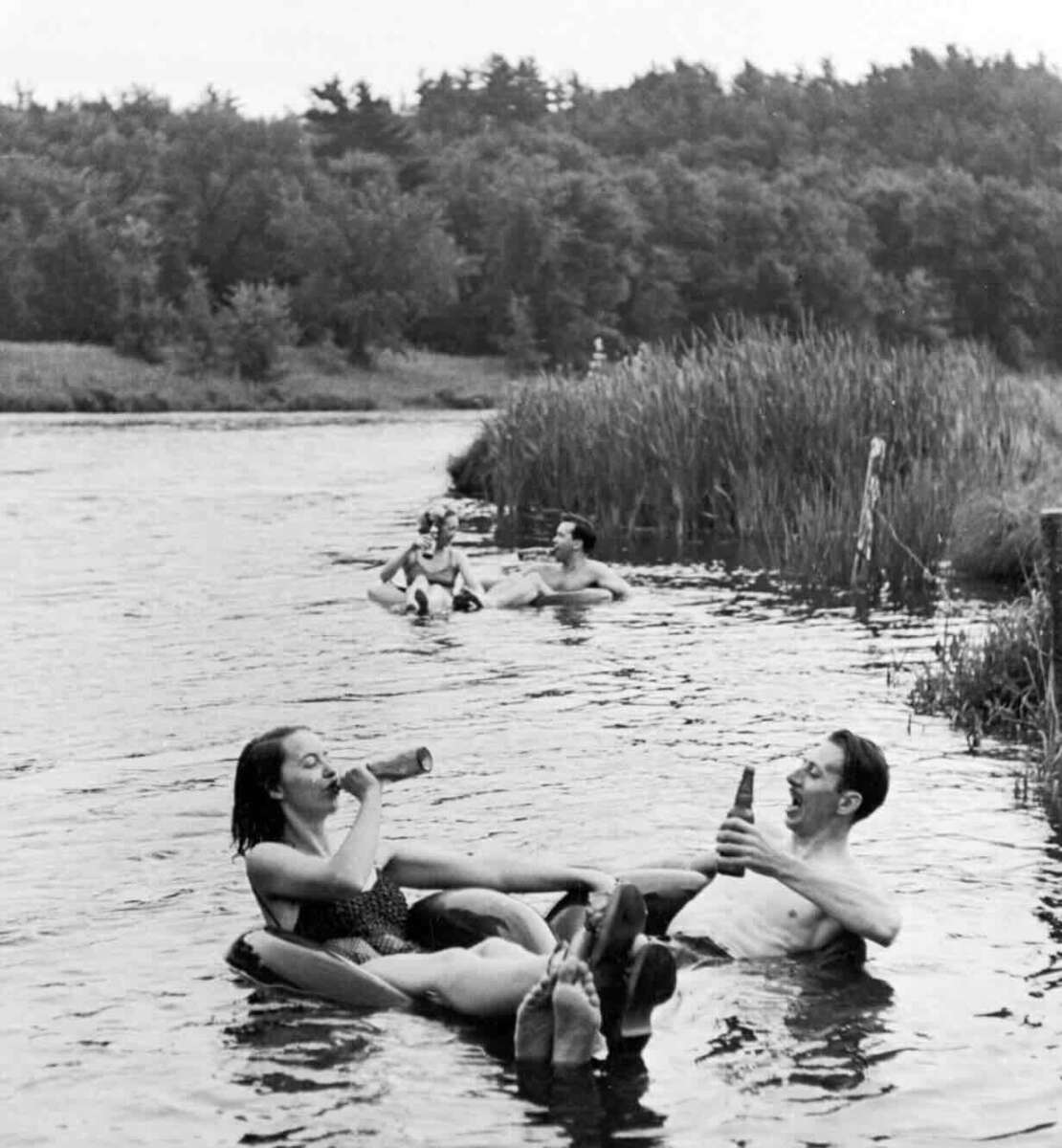 Four people float on inner tubes in a river, surrounded by trees and reeds. Two people in front are relaxing and drinking from bottles, while two more are seen further behind, also floating and enjoying the water.