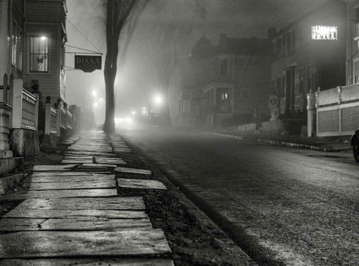 A foggy, empty street at night, lit by streetlights. Sidewalks line both sides, with old houses and a sign reading “DIANA” visible. The scene appears quiet and atmospheric.