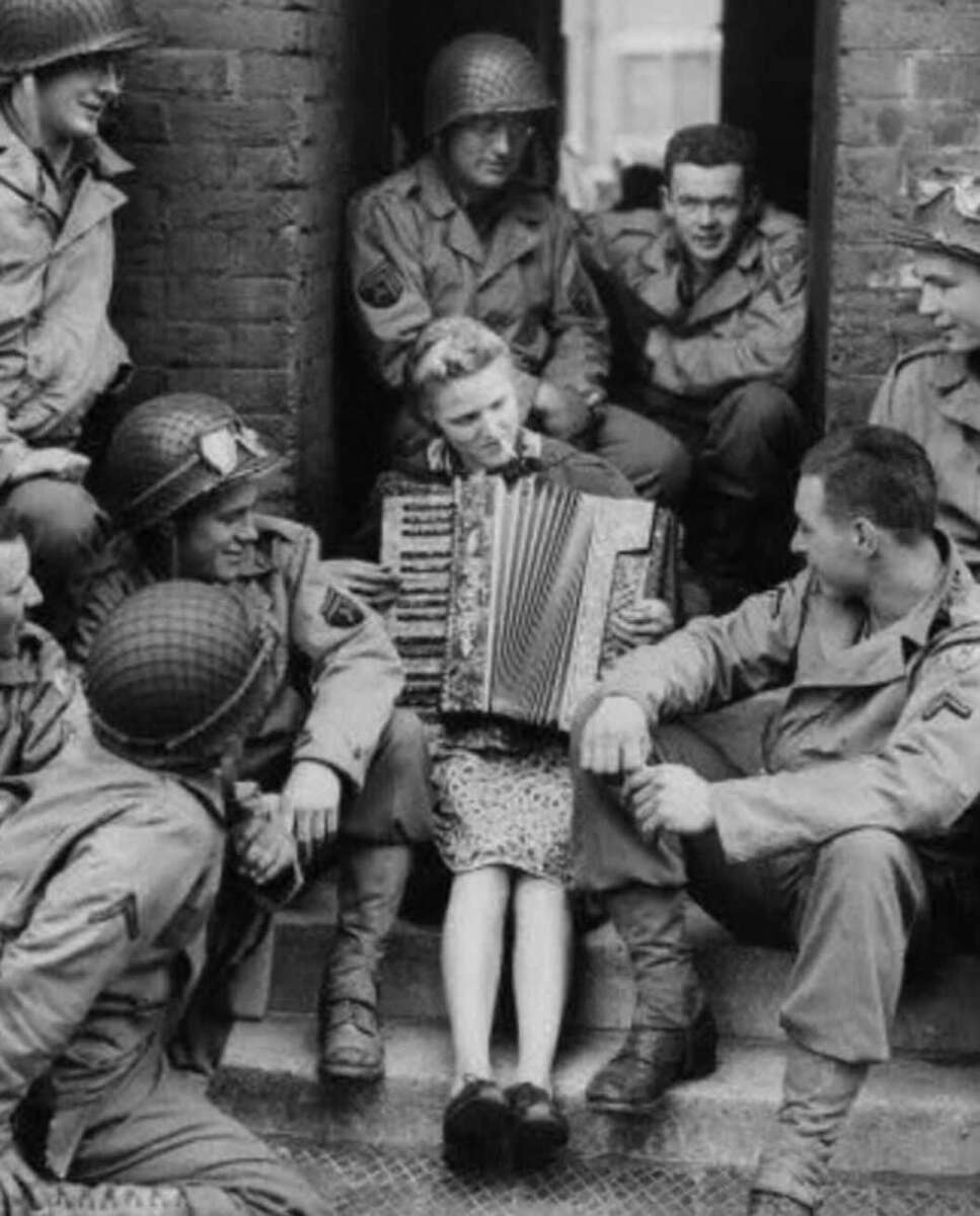 A woman sits in a doorway playing an accordion, surrounded by smiling soldiers in World War II-era uniforms and helmets, who are sitting and standing around her, listening intently.