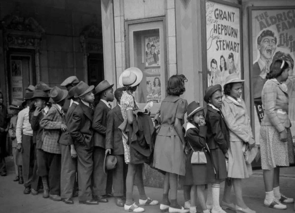 A group of children in vintage clothing and hats stand in line outside a movie theater, waiting to buy tickets. Movie posters are visible on the wall behind them.