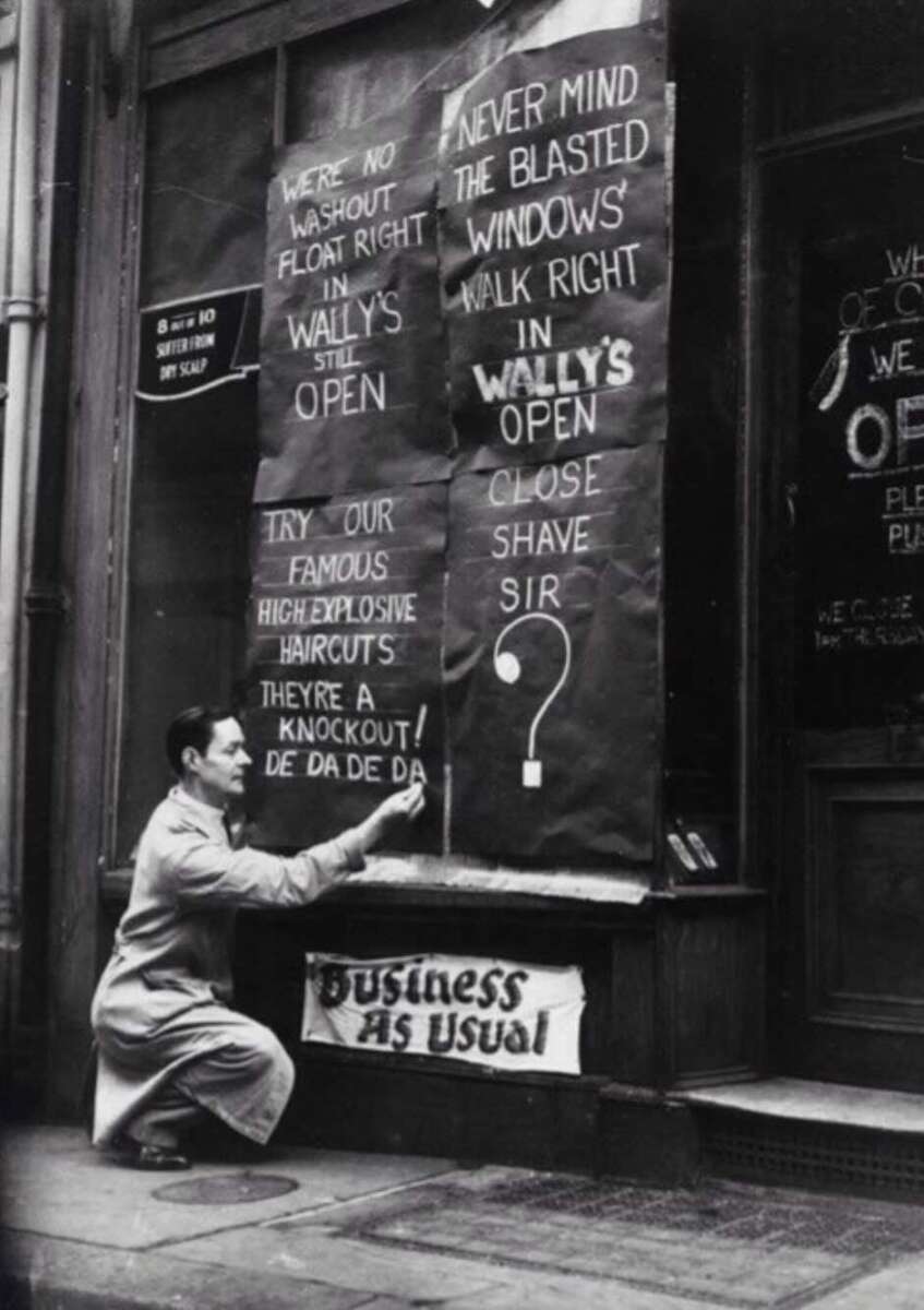 A man kneels outside a shop, pasting a sign with handwritten messages on the boarded-up windows, including “Wally’s still open” and “Business as usual,” despite visible damage to the storefront.