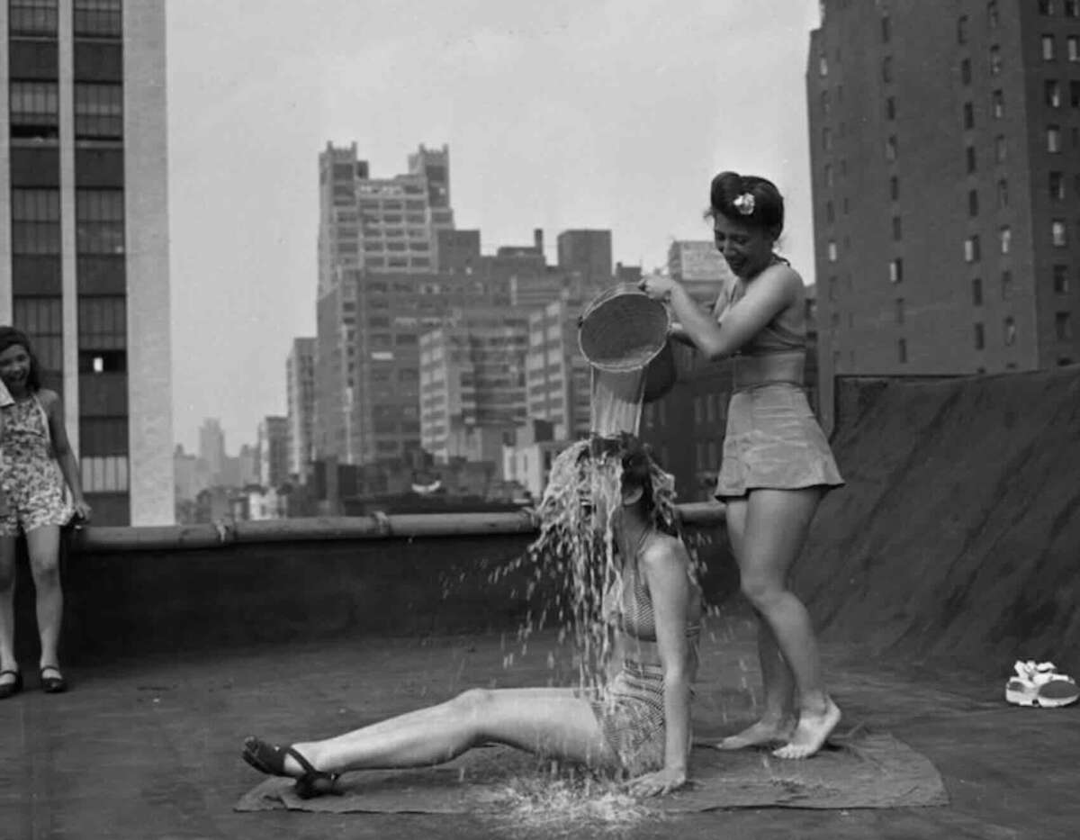 Two women in swimsuits are on a city rooftop. One is sitting on a mat while the other pours water over her from a bucket. Another woman stands nearby, watching. Tall buildings fill the background.