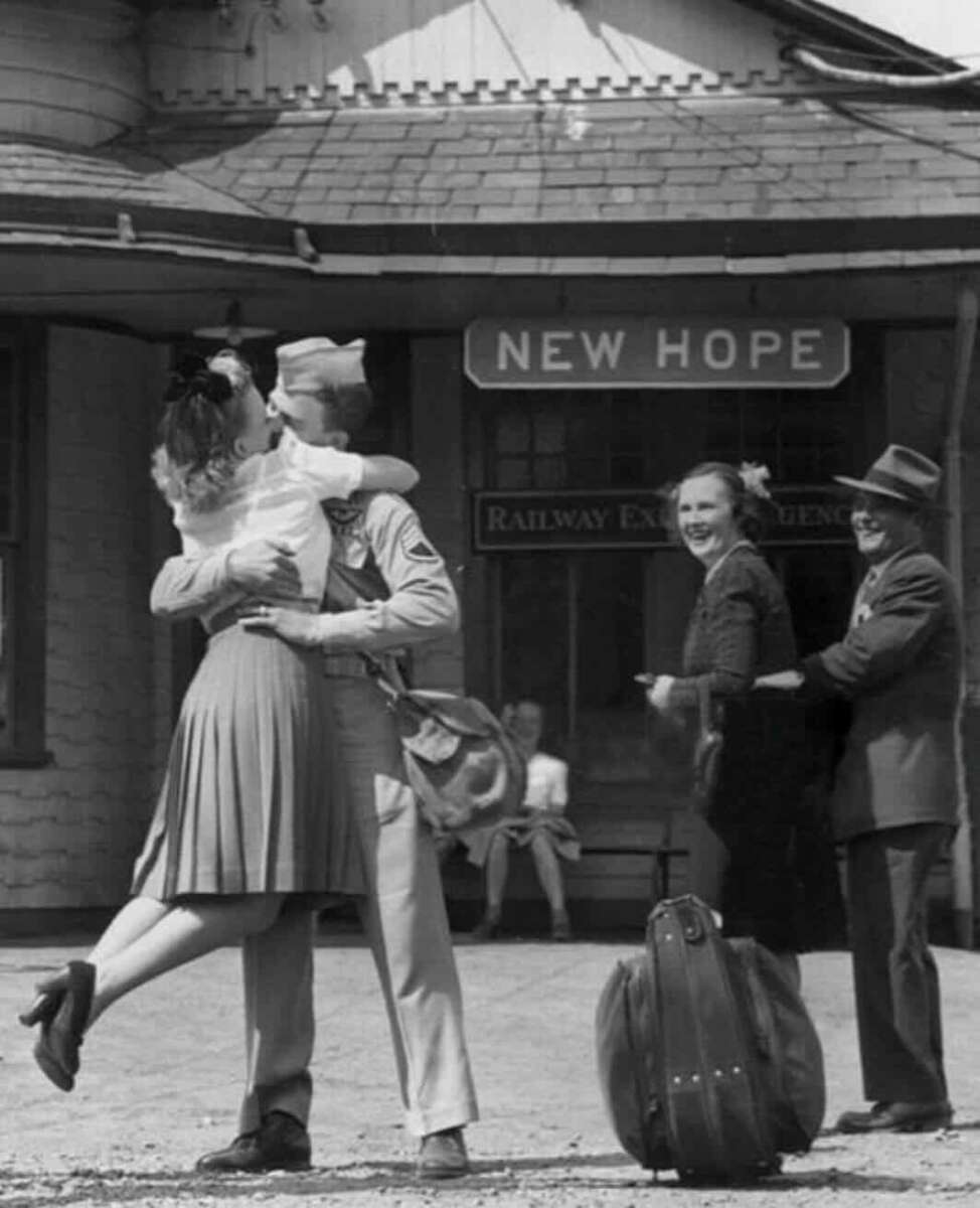 A soldier in uniform embraces and kisses a woman at a train station labeled "New Hope." Two smiling onlookers stand nearby, and luggage sits on the ground in the foreground.