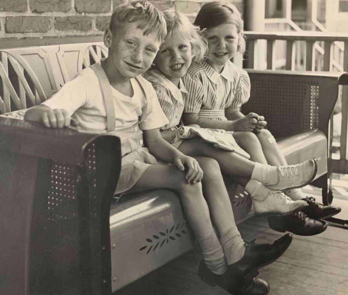 Three young children, two girls and a boy, sit closely together on a bench outside. They are smiling and dressed in vintage clothing, with the girls wearing dresses and the boy in suspenders and shorts.