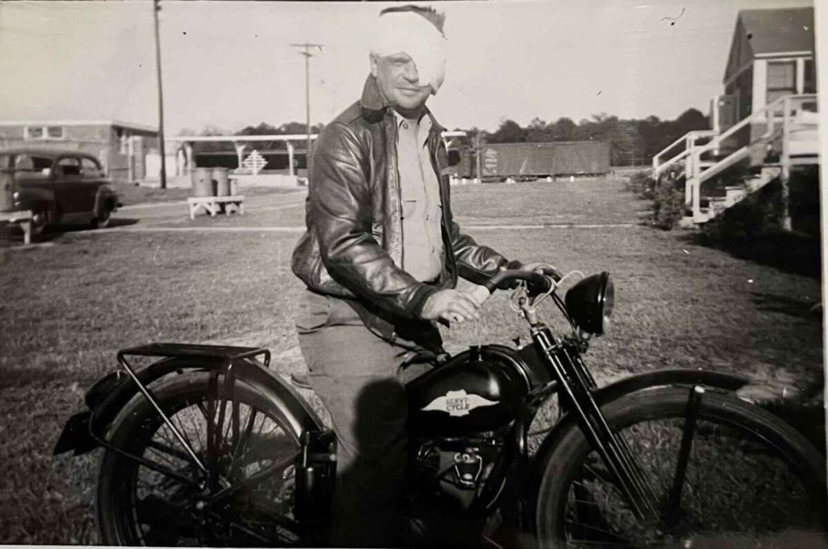 A man with a bandage over one eye sits on an Indian Chief motorcycle in an open grassy area, wearing a leather jacket. Buildings and vintage cars are visible in the background. The photo appears to be from the mid-20th century.
