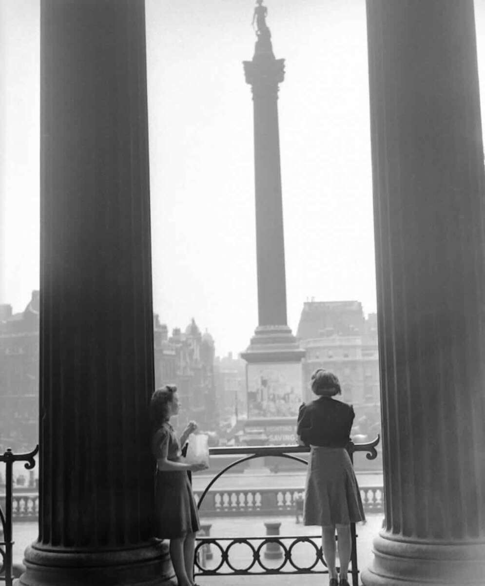 Two women stand on a balcony between large columns, looking out toward Nelson’s Column in Trafalgar Square, London. City buildings and the tall monument are visible in the background on a cloudy day.