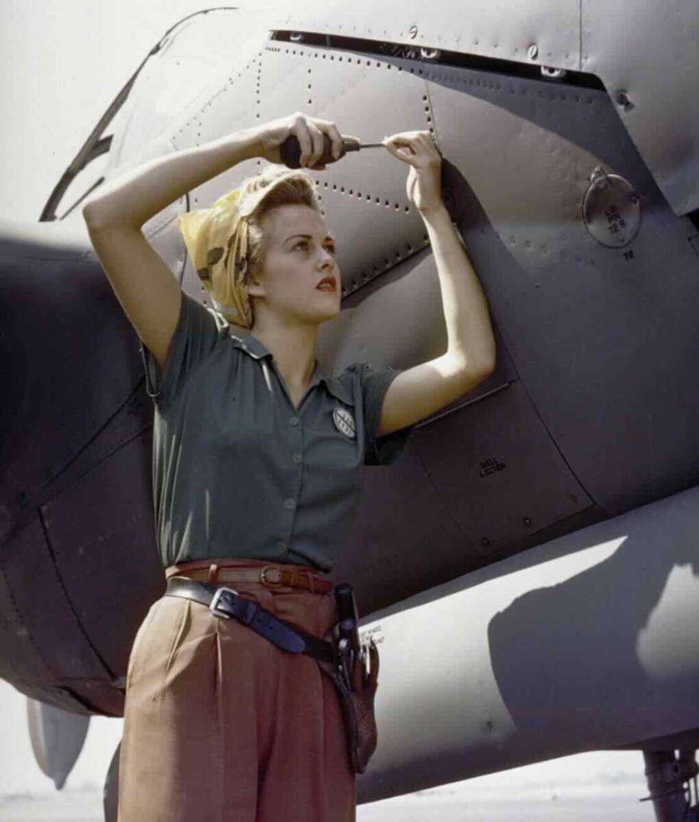 A woman in 1940s work clothes uses a screwdriver on an aircraft engine, wearing a yellow headscarf, short-sleeved green shirt, brown trousers, and a tool belt.