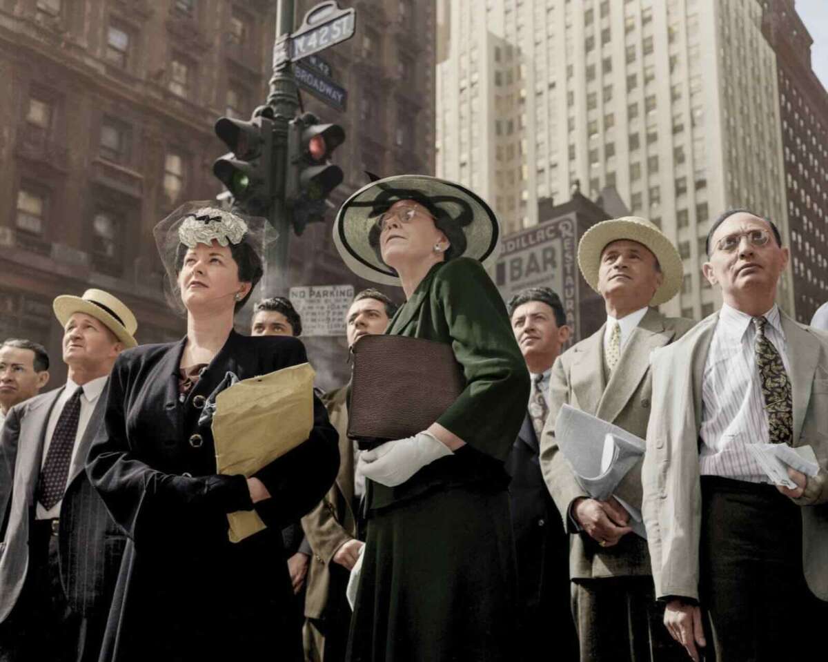 A group of men and women in 1940s attire stand on a city street, looking upward. Tall buildings and a traffic light are in the background, and the scene is bathed in daylight.