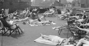 Black and white photo of people sunbathing on a city rooftop, lying on towels or sitting in lounge chairs, with tall buildings visible in the background.